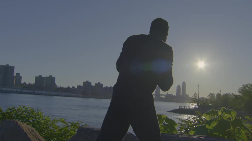 A silhouetted man performs slow-motion tai chi at sunrise with a city skyline view over the river