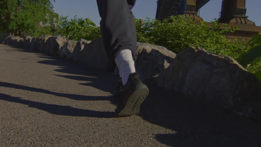 Low-angle slow-motion footage of a person's feet walking along a stone-lined path in a park with a bridge in the background