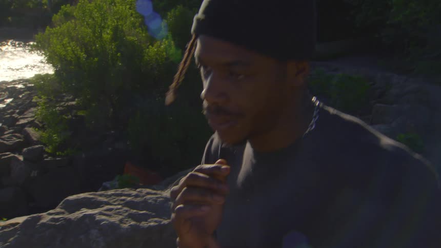 An expressive young man with dreadlocks freestyles and dances on a rocky riverbank in the late afternoon sun