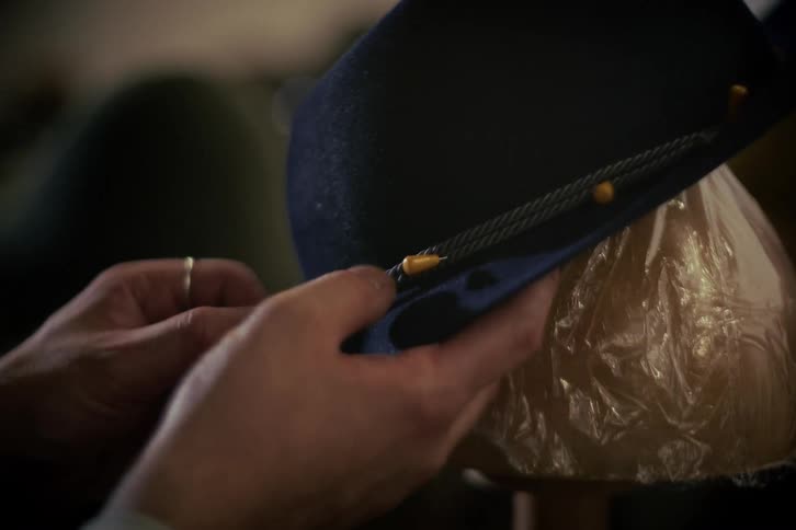 A milliner's hands carefully pin a decorative braided cord onto a classic dark blue felt hat