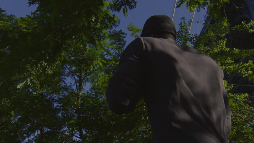 A slow-motion, low-angle shot from behind shows a man jogging in place under a bridge surrounded by trees