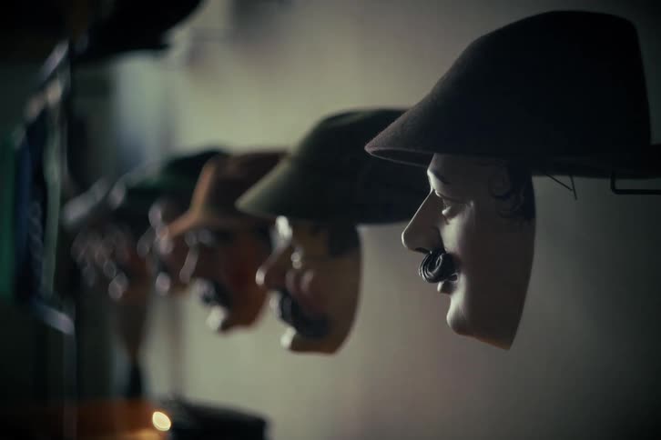 A creepy row of traditional carnival masks with hats are displayed on a dimly lit wall