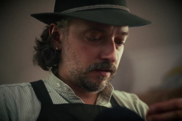 A skilled milliner in an apron and hat concentrates as he hand-sews a custom felt hat in his workshop