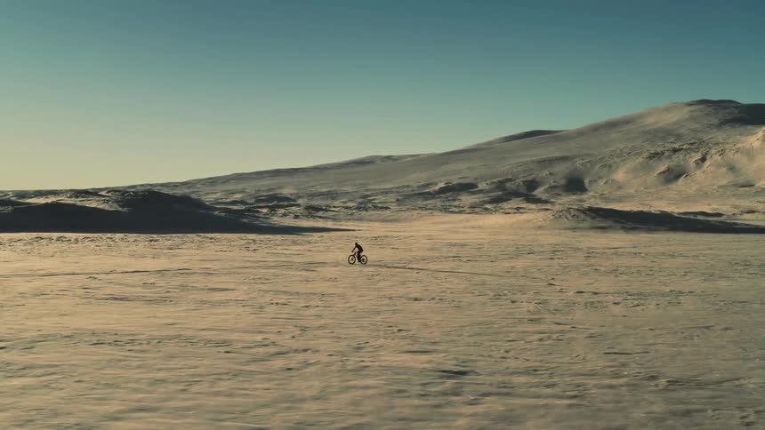 A lone cyclist pedals through an immense and desolate snow-covered landscape under a clear sky