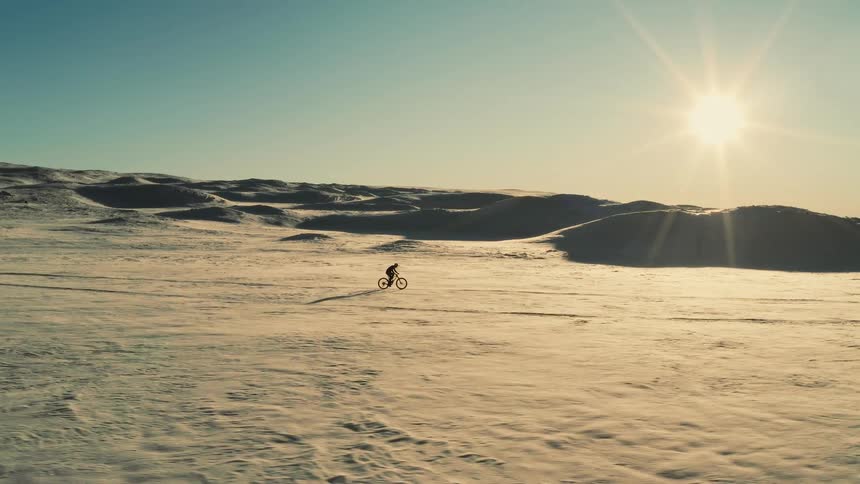 A lone cyclist on a fat bike traverses a vast, sun-drenched snowy landscape in an epic aerial view