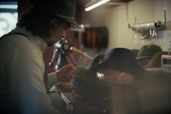 A skilled milliner hand-sews details onto a traditional felt hat in his atmospheric workshop