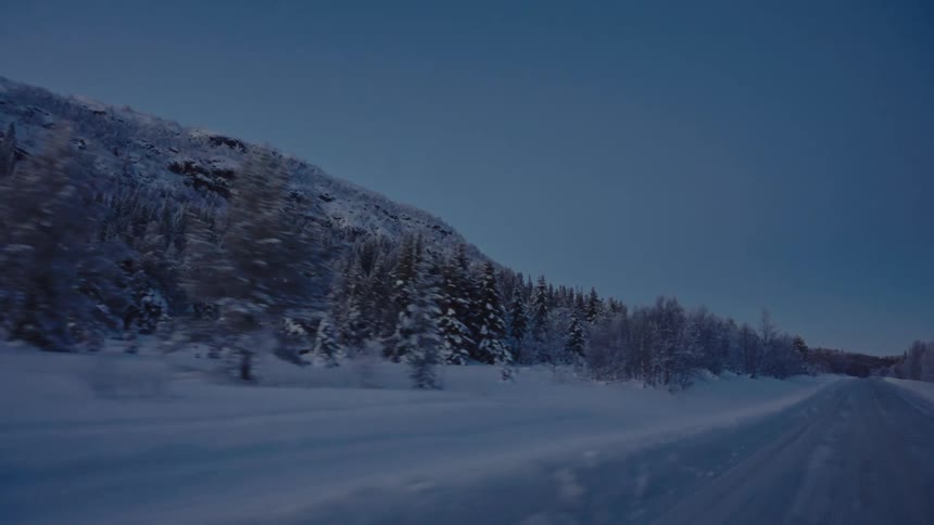 Point of view driving along a snowy road past a frozen forest and mountains at dusk