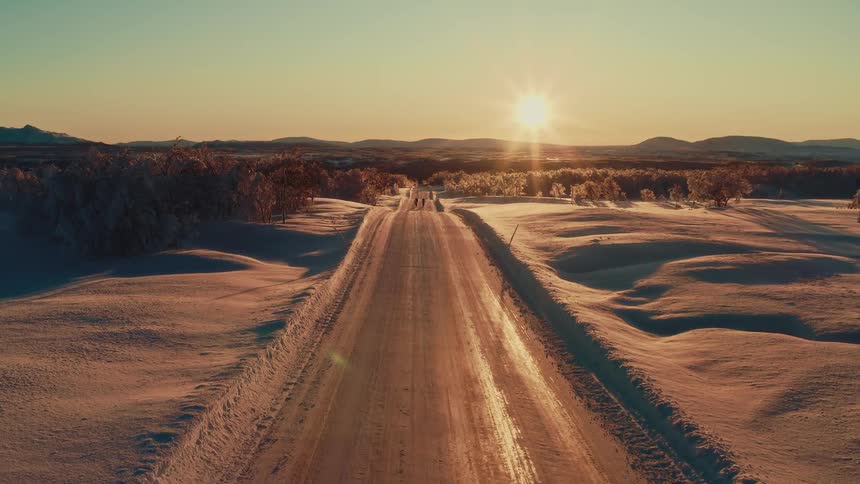 A stunning aerial view captures two people walking along a long, icy road through a vast snowy landscape at sunset