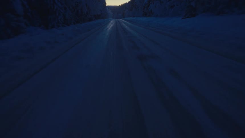 A low aerial view retreats down a desolate, snowy road flanked by a dark, frozen forest at twilight