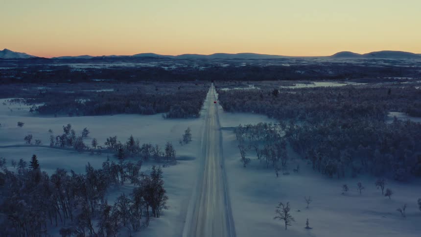 Aerial view of a long straight road running through a snow-covered arctic landscape towards the horizon at sunset