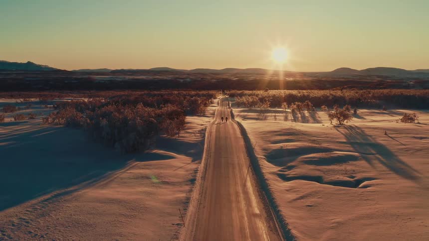 An awe-inspiring aerial view of two people walking along a remote, snowy road into the golden sunset
