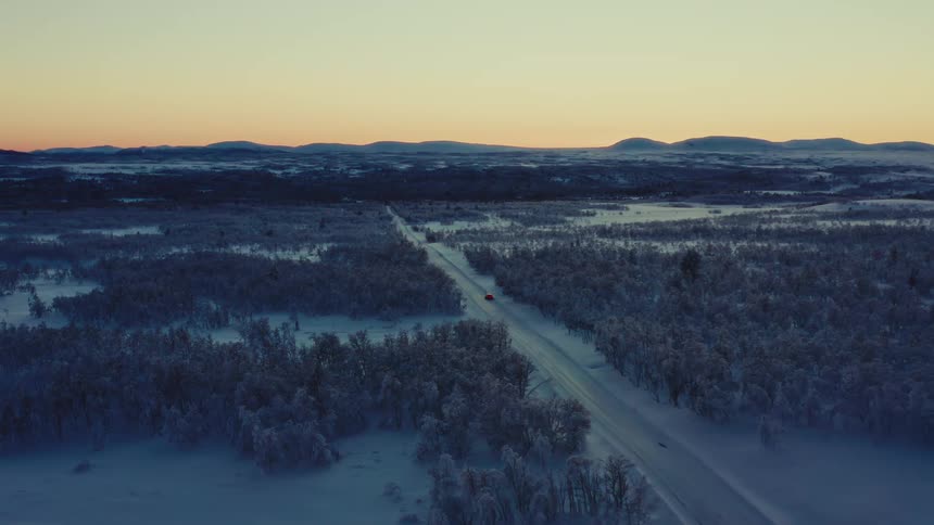 An aerial shot follows a red car driving along a snowy road that cuts through a vast, frozen forest landscape at sunset