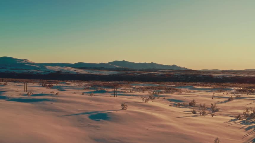 A serene aerial shot reveals a vast winter wonderland with snow-covered mountains bathed in the warm glow of the golden hour sun