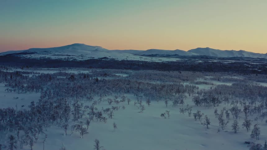 Aerial view pans slowly across a vast, snow-covered forest and mountain landscape during a tranquil winter sunset