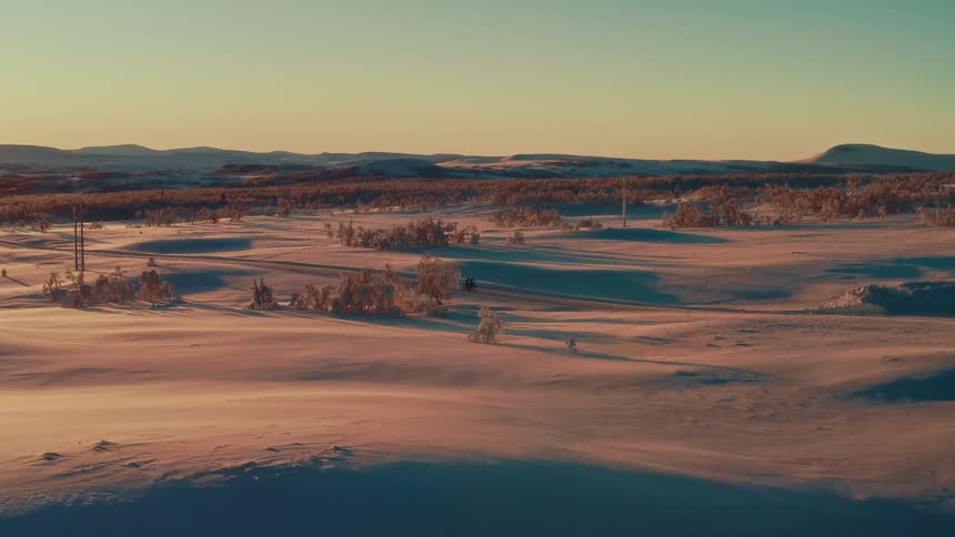 An epic aerial view follows two people riding fat bikes along a road through a vast, snowy landscape at golden hour