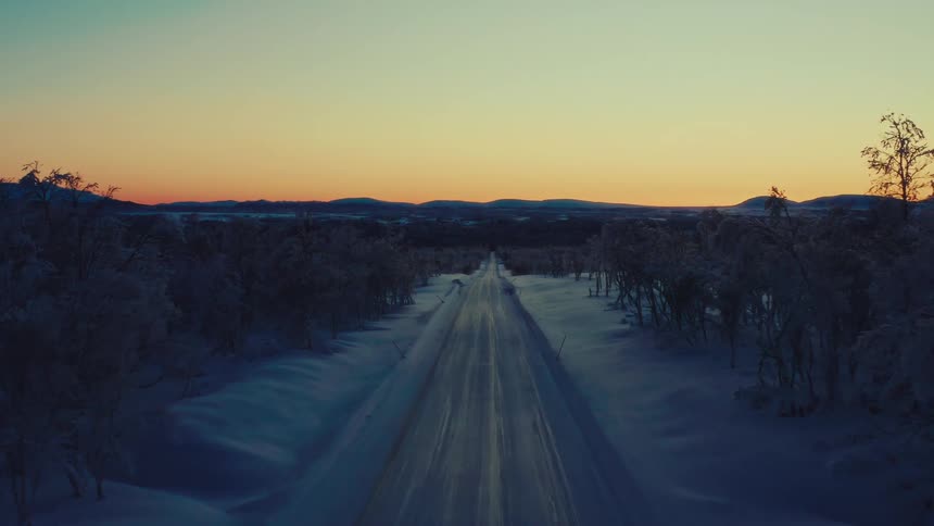 Cinematic aerial shot pulls back over a long, icy road stretching through a vast, snow-covered wilderness at sunset