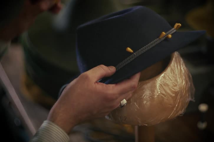A craftsman hand-sews a decorative band onto a custom blue fedora in a workshop