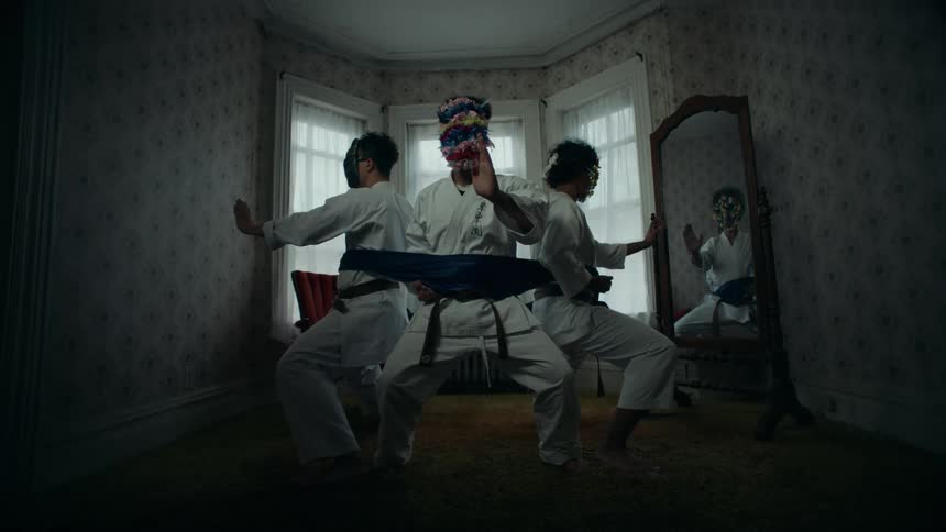 Three mysterious figures in karate gis and elaborate masks hold a synchronized stance while bound together by a single blue belt in a dimly lit room