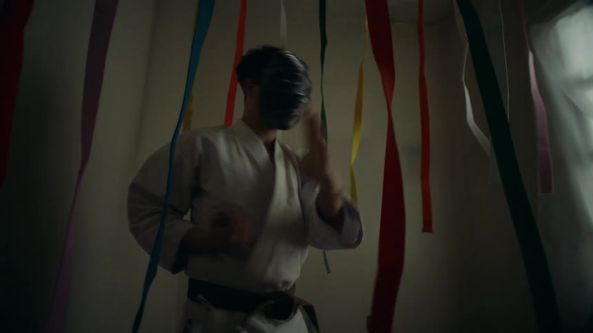 A mysterious masked man practices karate in a dark room filled with hanging colorful belts