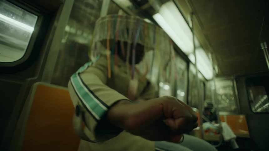 An artistic individual in a colorful fringed hat performs an expressive hand dance while sitting on a moving subway train