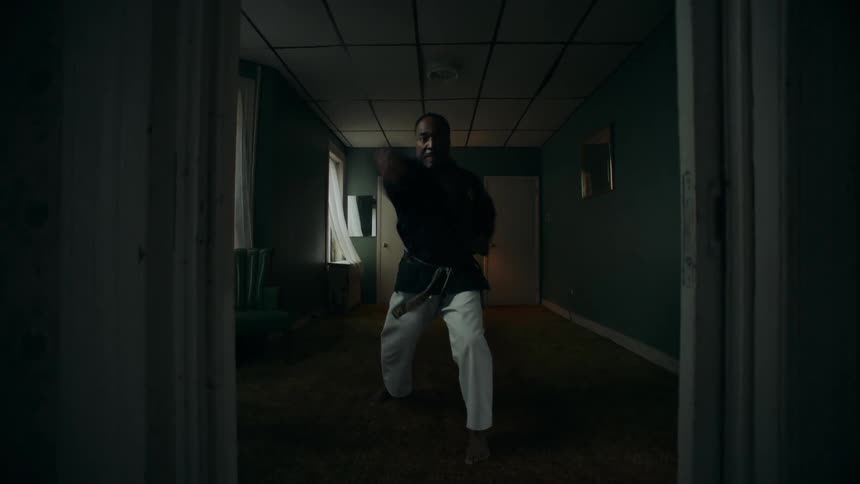 A man and woman practice martial arts in separate rooms of a dimly lit, vintage-style home