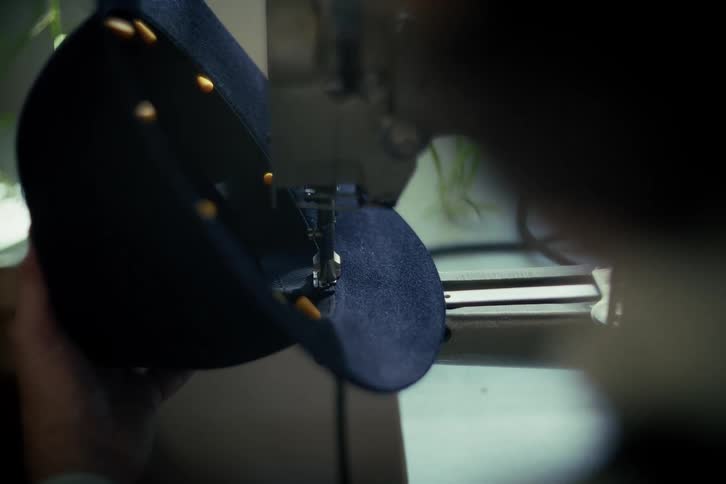 Over-the-shoulder close-up of a tailor's hands guiding denim fabric through a sewing machine to craft a cap