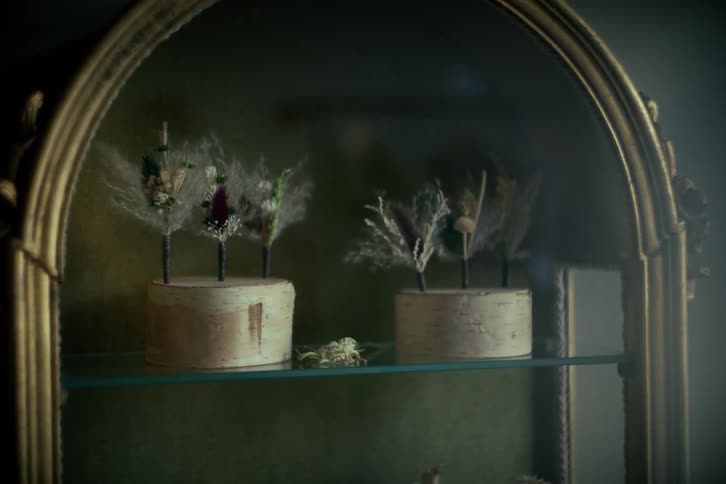Handmade dried flower boutonnieres are displayed on birch wood stands inside a vintage gold-framed cabinet