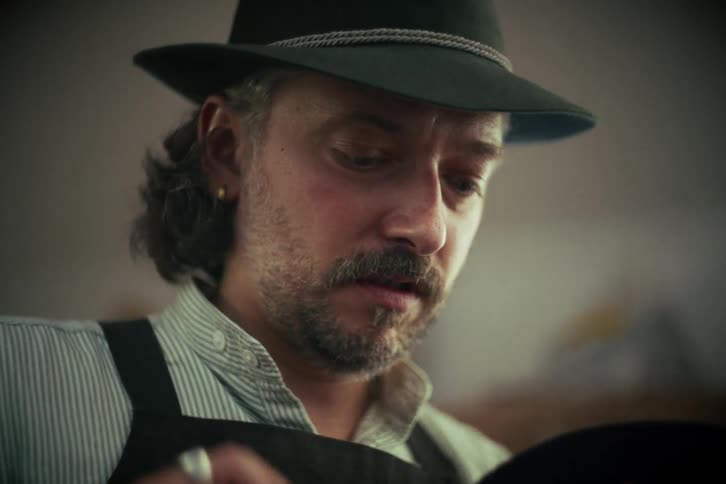 A skilled hatter with a mustache wearing a traditional hat concentrates as he crafts another hat in his workshop