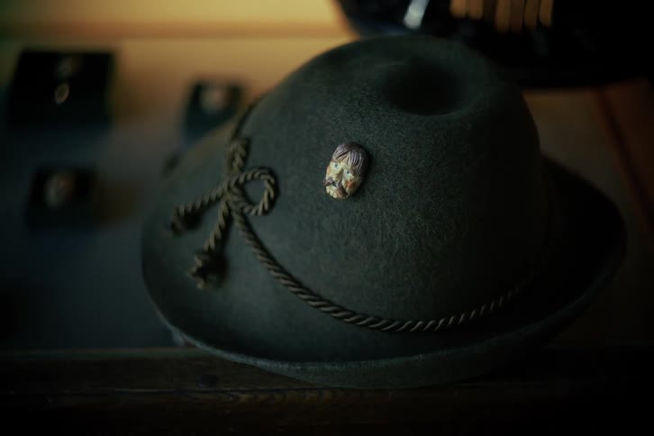 A close-up of a vintage-style, dark green felt hat with a decorative pin and rope band on display