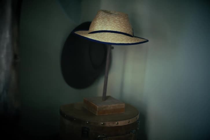 A vintage straw fedora with a blue band rests on a display stand atop an old trunk in a softly lit corner