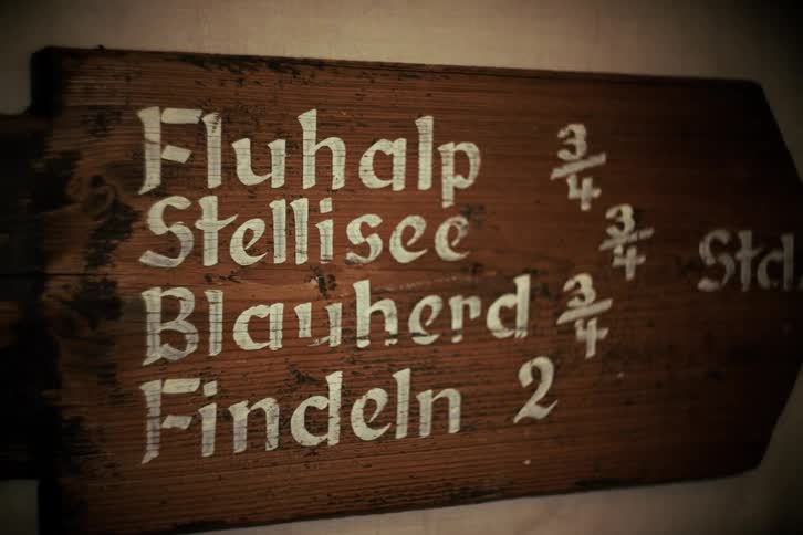 A close-up of a rustic, hand-painted wooden sign indicating hiking trail directions and times in German
