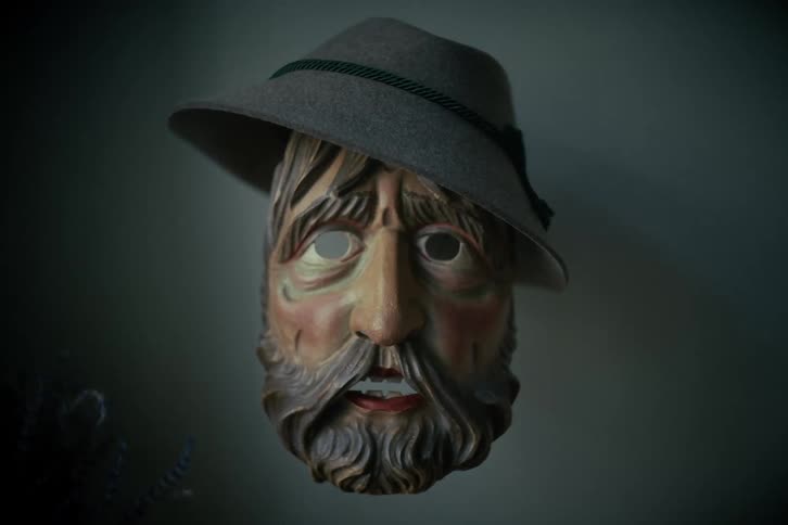 A traditional carved wooden mask of a bearded man in a Tyrolean hat hangs on a wall in moody, dim lighting