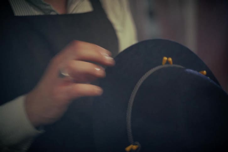 A skilled hatter in an apron meticulously hand-sews a decorative band onto a classic felt hat