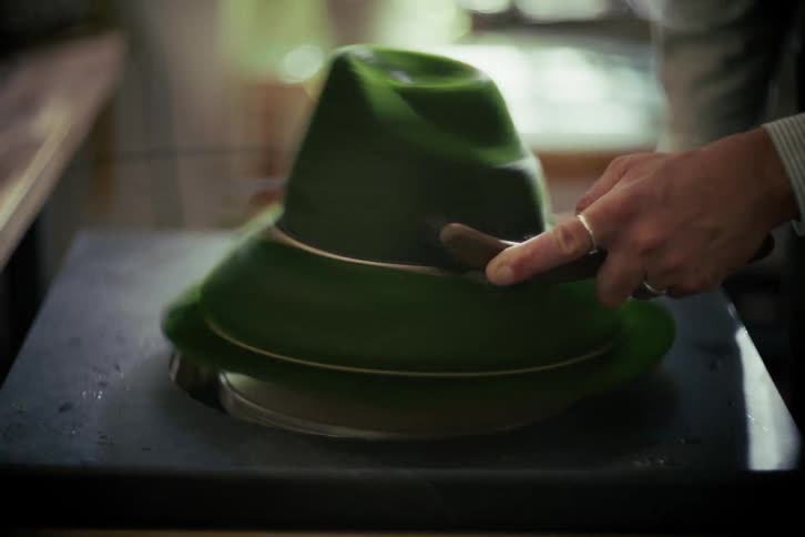 A skilled milliner carefully brushes a green felt fedora as it spins on a shaping machine in a workshop