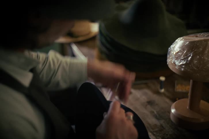 A master hatter meticulously hand-sews a decorative cord onto a felt hat in his traditional workshop
