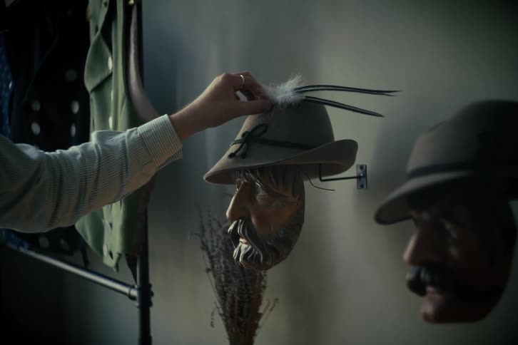 A person's hand carefully arranges feathers on a traditional alpine hat resting on a carved wooden mask