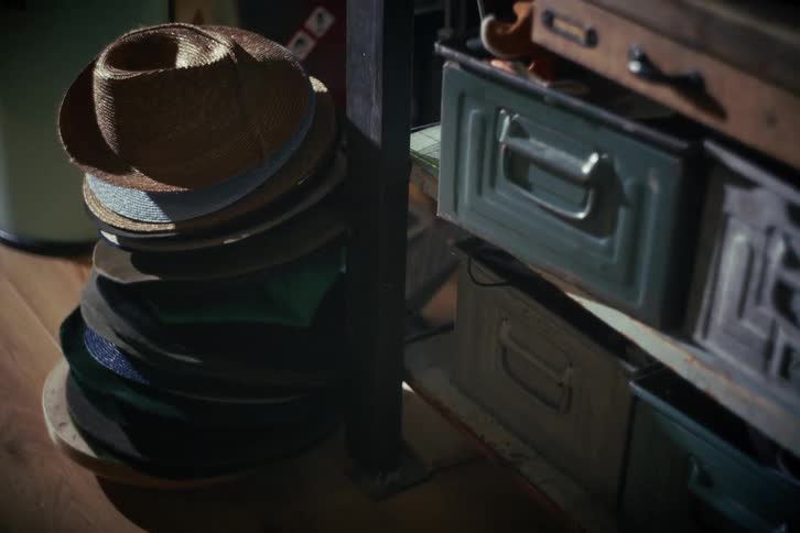 A stack of vintage fedora hats sits on a wooden floor next to old metal drawers in a dimly lit, atmospheric room