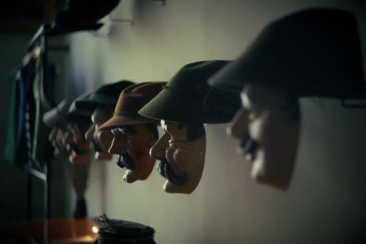 A cinematic rack focus shot reveals a row of traditional carnival masks with hats hanging on a dimly lit wall