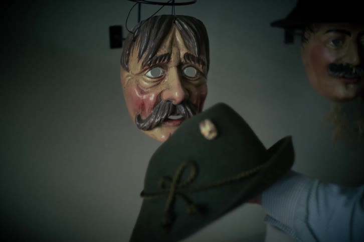 A hand holds a traditional green felt hat with unsettling vintage carnival masks hanging in the background