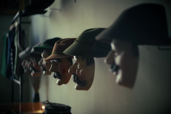 A cinematic rack focus reveals a line of traditional character masks with hats hanging on a wall in a dark room