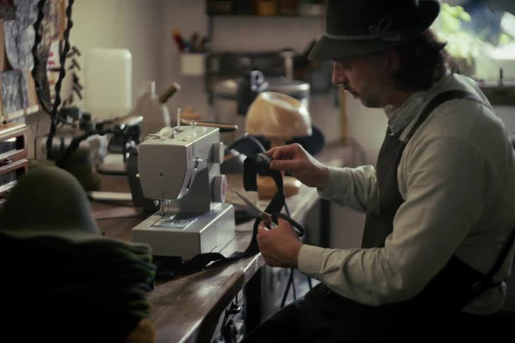 A traditional hat maker wearing an apron and hat sits at his workbench, using a sewing machine to meticulously craft a hat band