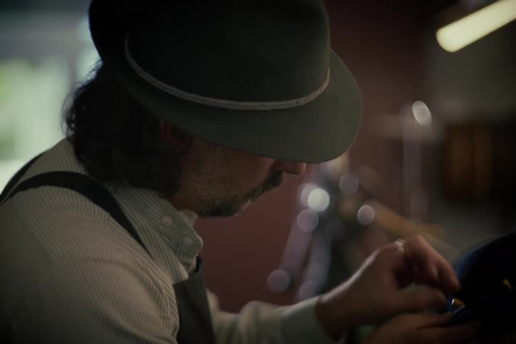 A craftsman wearing a hat and an apron carefully sews a piece of fabric by hand in his workshop