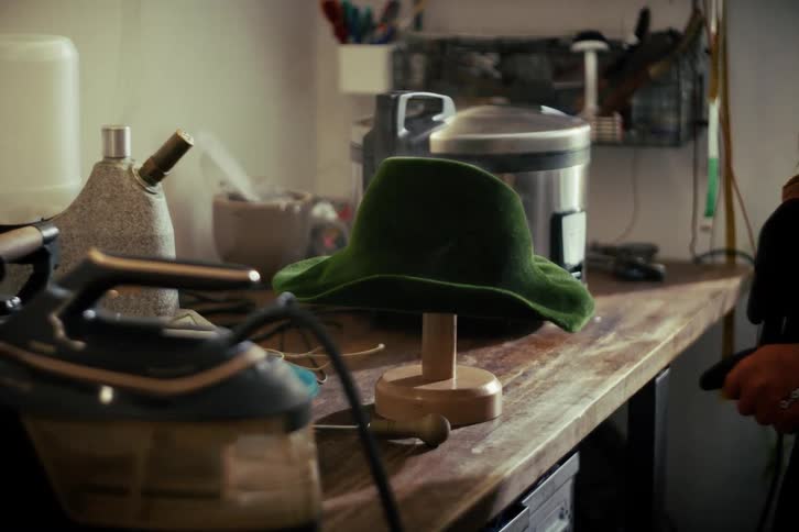 An artisan steams a new handmade green felt hat on a wooden stand in a workshop