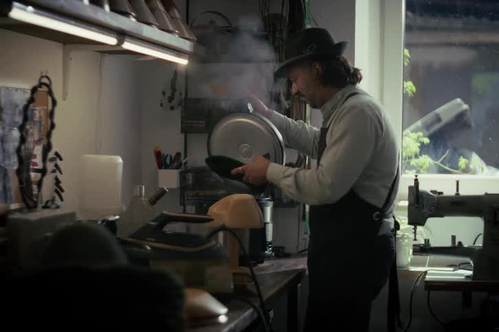 A skilled milliner steams a dark green felt hat body over a steamer in his workshop, preparing it for shaping