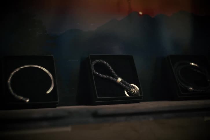 A stylish collection of men's silver and leather bracelets and a keychain are elegantly displayed in black boxes with a moody, artistic backdrop