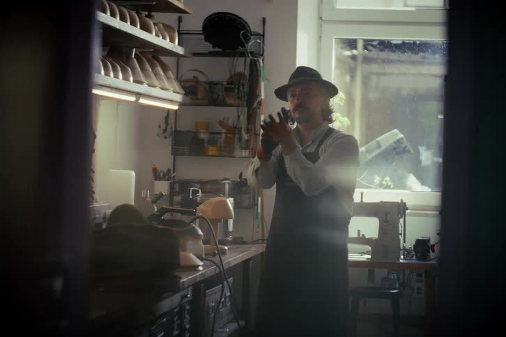 A mustachioed artisan in a hat and apron carefully inspects leather gloves in his vintage workshop