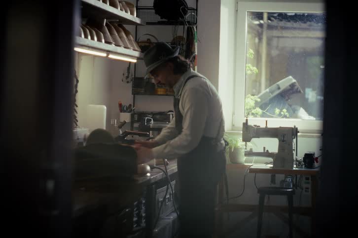 A traditional hat maker carefully steams and shapes a hat on a wooden form in his sunlit artisan workshop