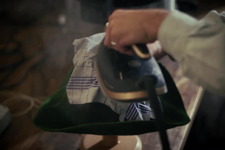 A craftsman carefully irons a green felt hat using steam to shape it in a workshop