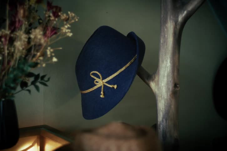 A classic blue Tyrolean hat with a golden cord hangs on a rustic wooden hat rack next to a vase of dried flowers