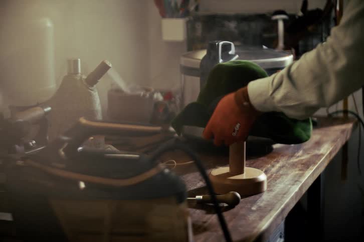 A milliner steams a green felt hat form and skillfully shapes it on a wooden block in a hazy workshop